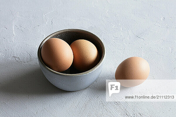 Studio still life of a bowl with three brown eggs and a single egg on a textured surface.