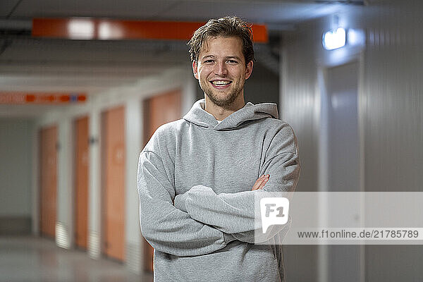 A smiling man in a casual gray hoodie crosses his arms while standing in a brightly lit hallway with orange doors and a modern  industrial design  Molenhoek  Netherlands