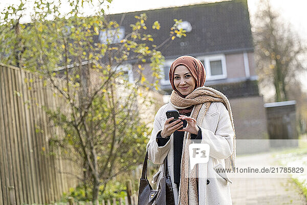 Smiling woman in a hijab holding a smartphone outdoors on a sunny day  Netherlands