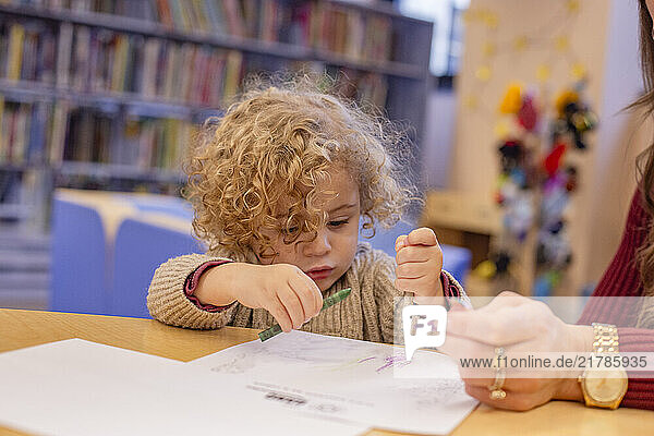 Curly-haired child focused on drawing with crayons at a table in a library while a person sits beside them. Cleveland  Ohio  USA