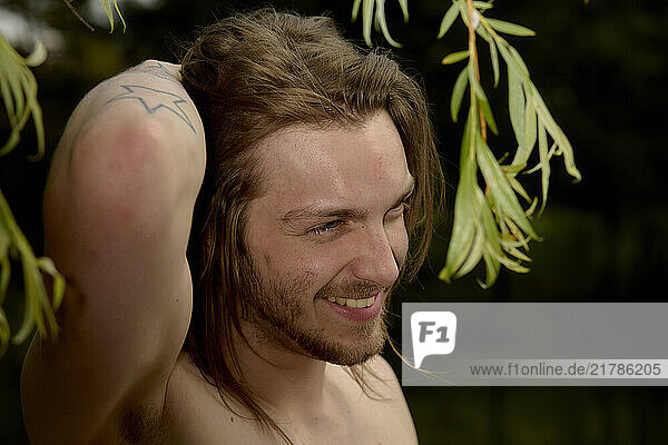 Smiling man with long hair and tattoos standing outdoors with green foliage in the background