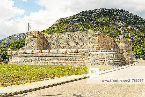 A stone fort of Kastio with battlements and flags under a cloudy sky  flanked by a green mountain and roadside  Ston Croatia