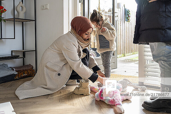 Mother in a hijab is helping a child put on shoes by the front door as another child waits beside them  Netherlands