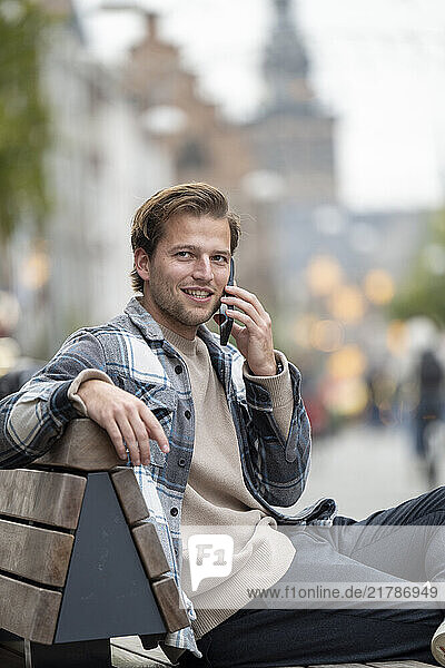 A young man with short hair  wearing a plaid shirt and beige sweater  is sitting on an outdoor bench while talking on his smartphone  smiling naturally  Nijmegen  Netherlands