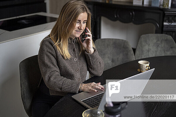 A woman with long blonde hair wearing a brown sweater is sitting at a black table  talking on a cellphone  and using a laptop. Netherlands