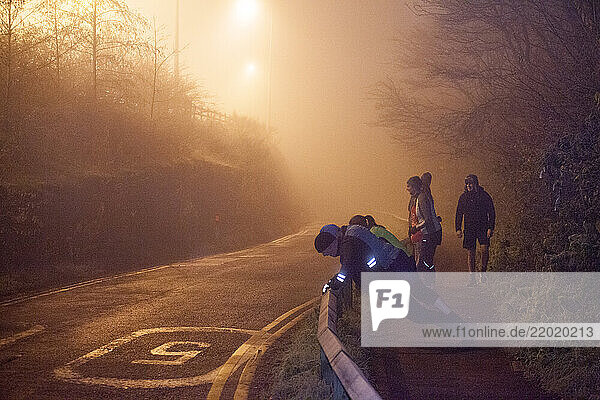 A group of people exercise outdoors in a misty  dimly lit street at dusk  illuminated by streetlights  Sale Water Park  Manchester  UK