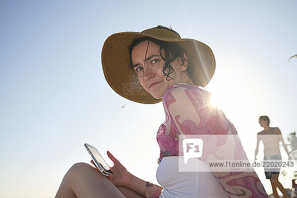 Woman in sun hat holding phone on beach with sunlight behind  man walks in background  concerned  Florida Keys  USA