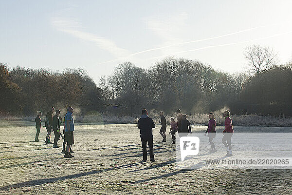 A group of people dressed in athletic gear perform exercises on a frost-covered field surrounded by trees  Sale Water Park  Manchester  UK