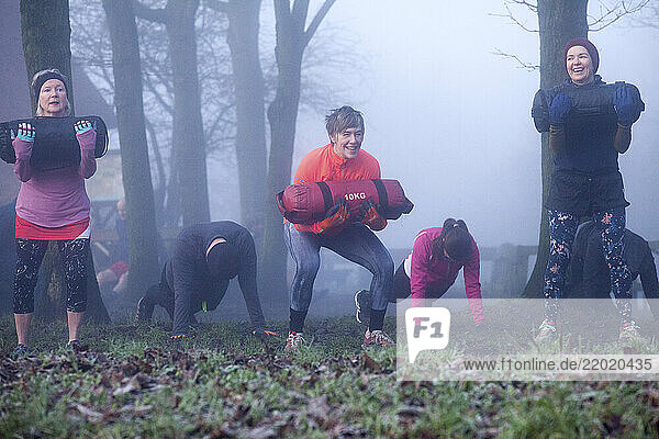 A group of adults exercising outdoors in a misty forest  using weighted sandbags and wearing colorful athletic clothing. Sale Water Park  Manchester  UK