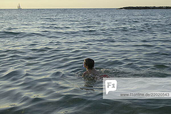Child swimming in the sea at sunset with a distant sailboat on the horizon. Israel