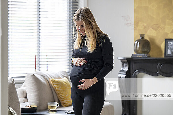 Pregnant woman in a cozy living room  gently cradling her belly while standing beside a modern fireplace. Netherlands