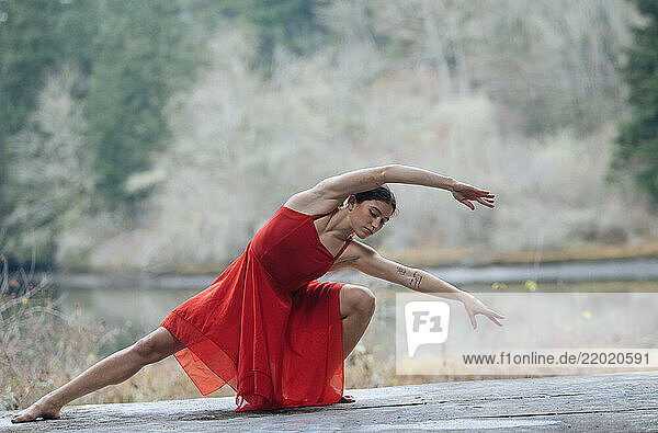 Woman in a red dress performing a graceful dance pose outdoors by a serene lake. USA
