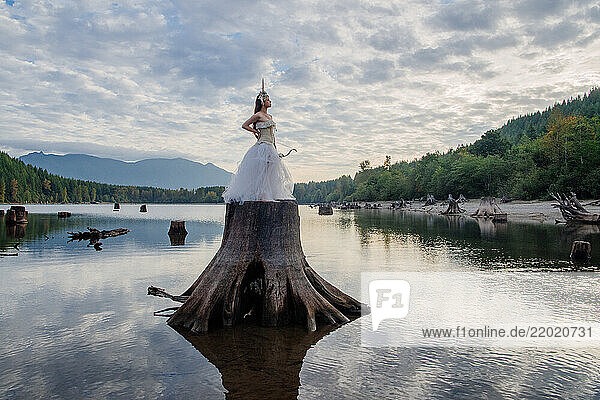 Woman with a bow in a wedding dress stands on a large tree stump in a serene lake surrounded by mountains. Rattlesnake Lake  Puget Sound  WA  USA