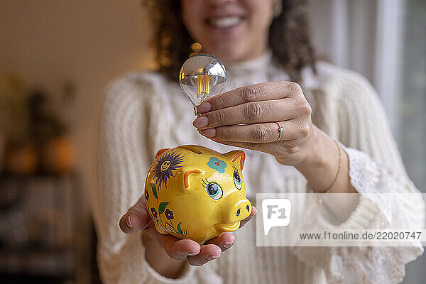 Woman holds a yellow floral piggy bank and a silver LED light bulb indoors. Netherlands
