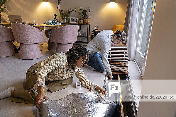 Couple installing insulation in a modern living room with pink chairs and decorative plants. Netherlands