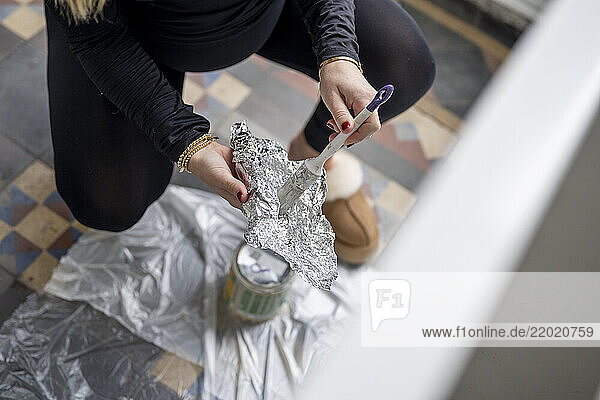 A person wearing a black outfit and fluffy slippers prepares a paintbrush wrapped in aluminum foil over a paint can on a tile floor. Netherlands