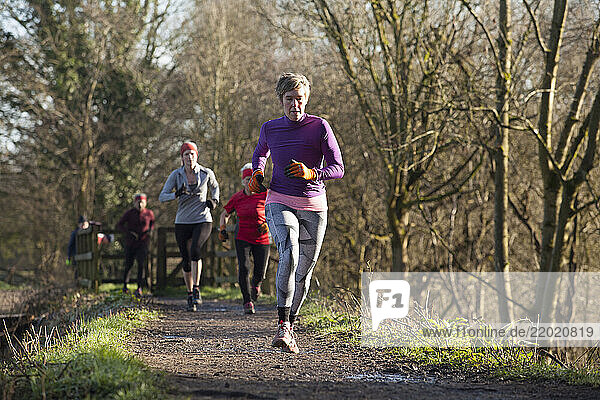 A group of runners jog along a sunny forest trail dressed in winter athletic wear  with leafless trees lining the path. Sale Water Park  Manchester  UK