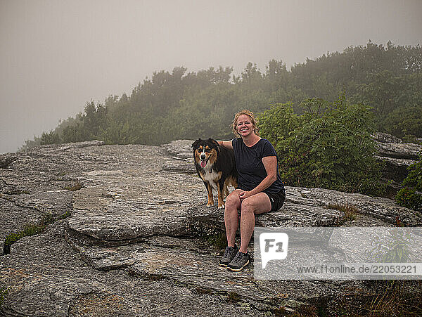 Portrait of smiling woman with Black Tri Australian Shepard sitting on rocks