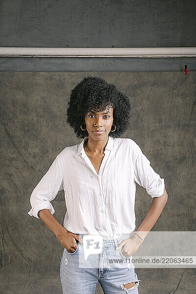 Smiling young woman standing with hands in pockets in front of gray colored backdrop