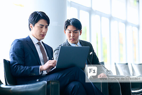 Two Japanese Businessmen Sitting On Chairs With A Laptop