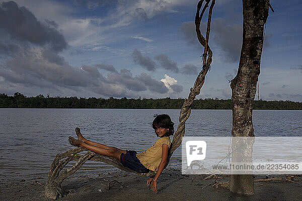Girl reclines on tree roots beside the river at the Kalabogi village in Khulna  Bangladesh  on 04 September 2023. In the coastal areas of Southern Bangladesh like Kalabogi  people‘s livelihoods depend entirely on nature. Climate change has severe impacts on the natural environment. The consequences range from rising sea levels leading to salinization of soils and groundwater to erratic rainfall patterns and more frequent and destructive tropical cyclones. People living in coastal areas fight for their survival every day; Khulna  Dhaka  Bangladesh