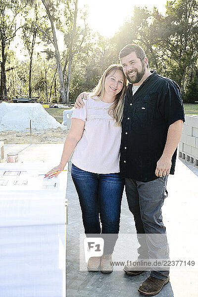 Smiling couple embraces at their home construction site with blueprints.
