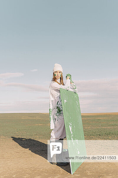 Young woman holding green painting on dry field