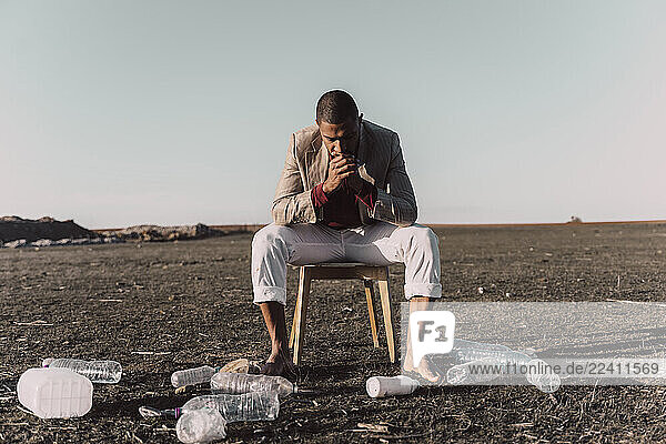 Young man sitting on chair surrounded by plastic bottles in barren land
