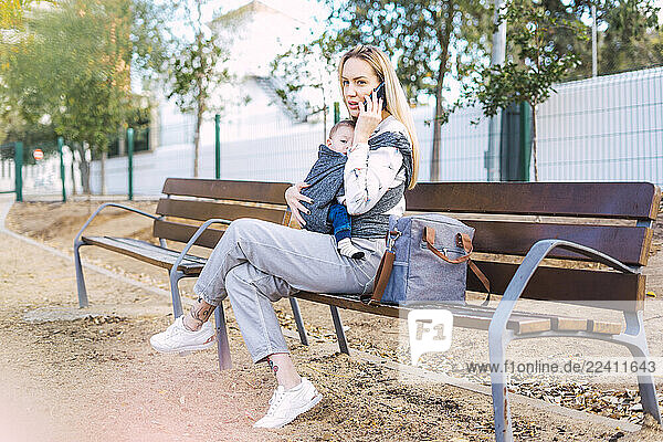 Mother resting with baby boy on a park bench talking on the phone