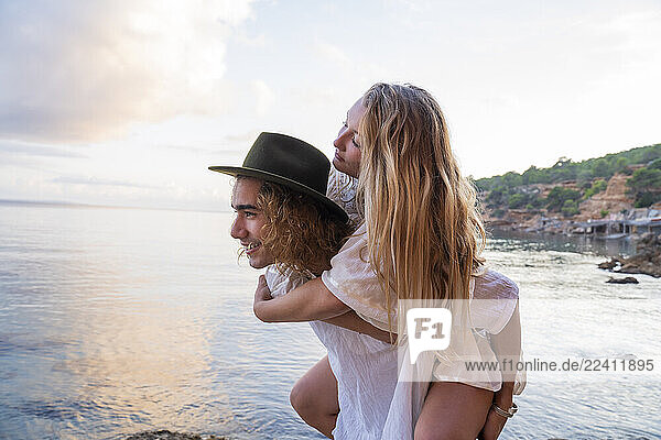 Young man giving his girlfriend a piggyback ride in front of the sea  Ibiza  Balearic Islands  Spain