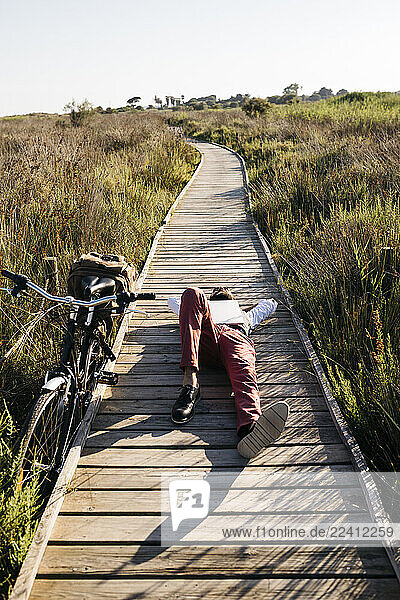 Well dressed man with laptop lying on a wooden walkway in the countryside next to a bike