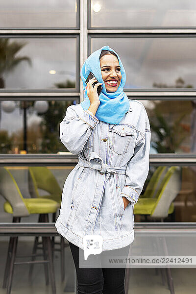 Young woman wearing turquoise hijab and phoning in front of a cafe
