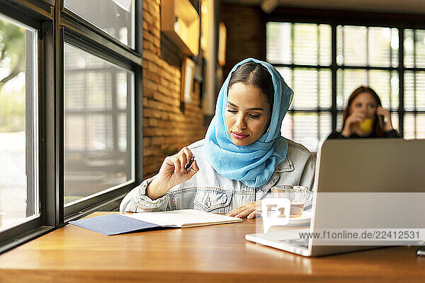 Businesswoman wearing turquoise hijab in a cafe and writing in notebook