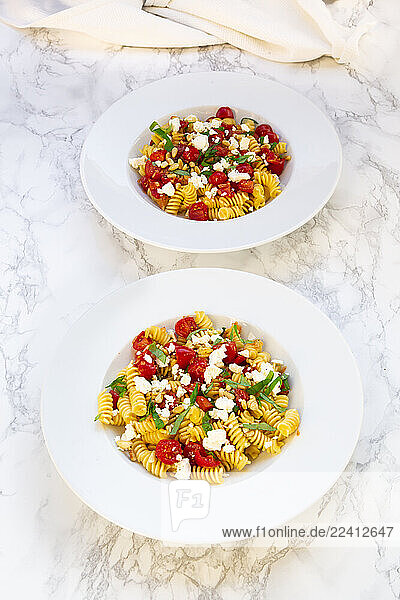 Two plates of pasta with tomatoes  feta cheese  and pine nuts on a marble background