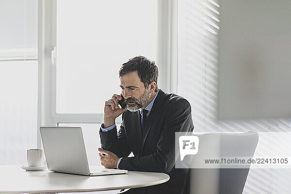 Mature businessman using laptop and cell phone on table in office