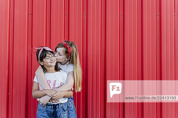 Two sisters standing in front of a red wall  kissing