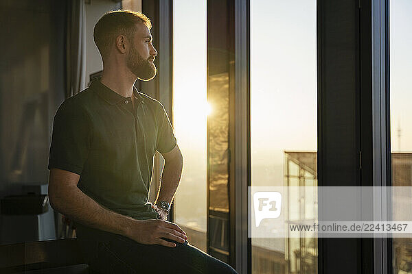Young man in modern office looking out of a glass window at sunset