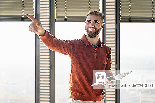 Young man smiling and pointing while holding a tablet in a modern office
