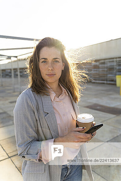 Portrait of young woman with coffee to go and mobile phone at backlight