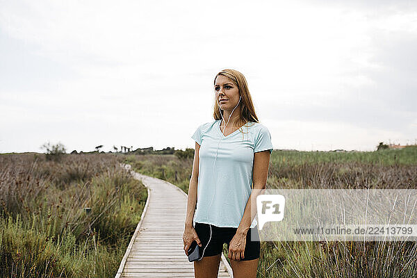 Female jogger with earphones and smartphone standing on a wooden walkway