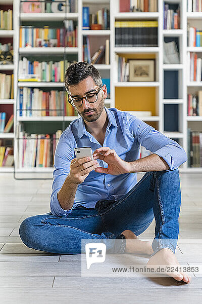 Portrait of smiling young man sitting in front of bookshelves on the floor at home using smartphone