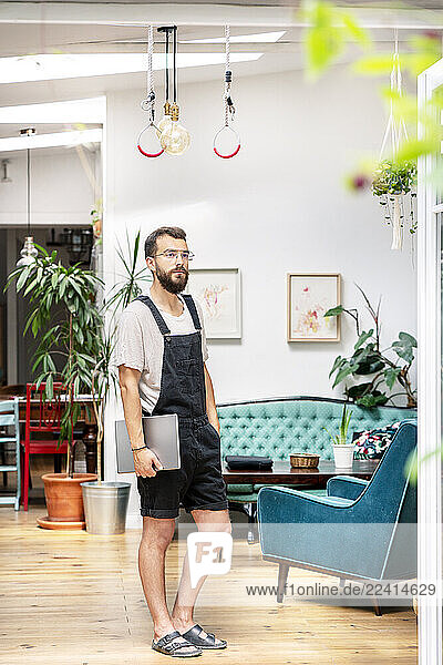 Young man in dungarees standing in his livingroom  holding digital tablet