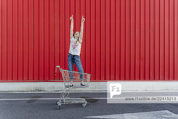 Girl in a shopping cart pointing fingers up in front of red wall