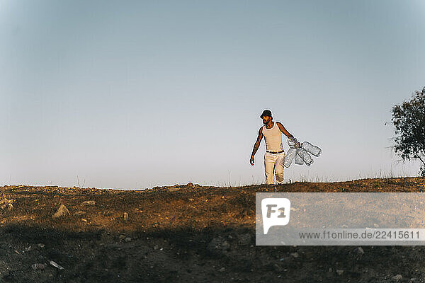 Young man walking in barren land holding empty plastic bottles