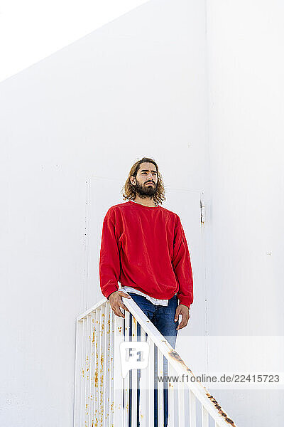 Portrait of bearded young man wearing red sweatshirt standing on stairs looking at distance