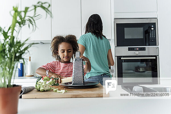 Girl cooking with mother in kitchen grating cheese
