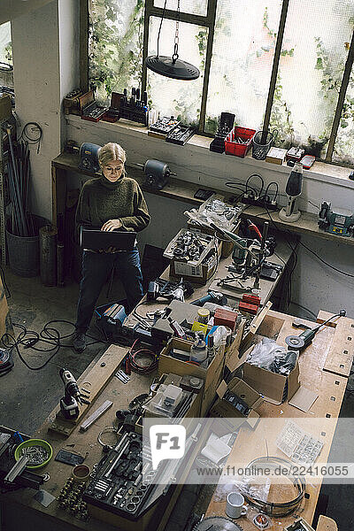 Small business owner working on a laptop in a workshop surrounded by tools and electronics