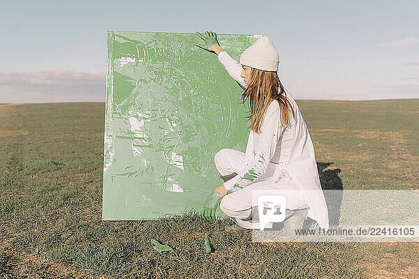 Young woman holding green painting  crouching on dry field