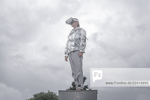 Girl in silver suit looking through VR goggles  standing on box  against grey sky