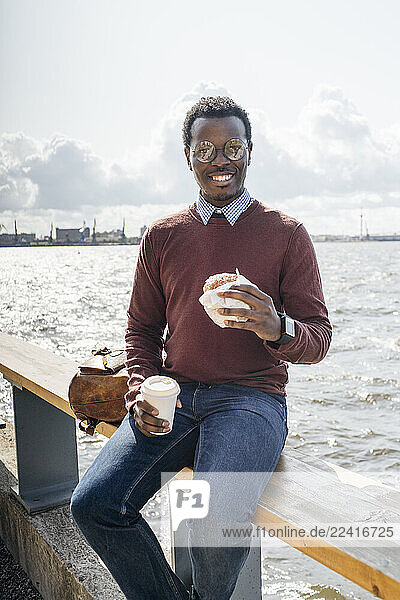Young man sitting on railing at the sea  eating hamburger  drinking coffee